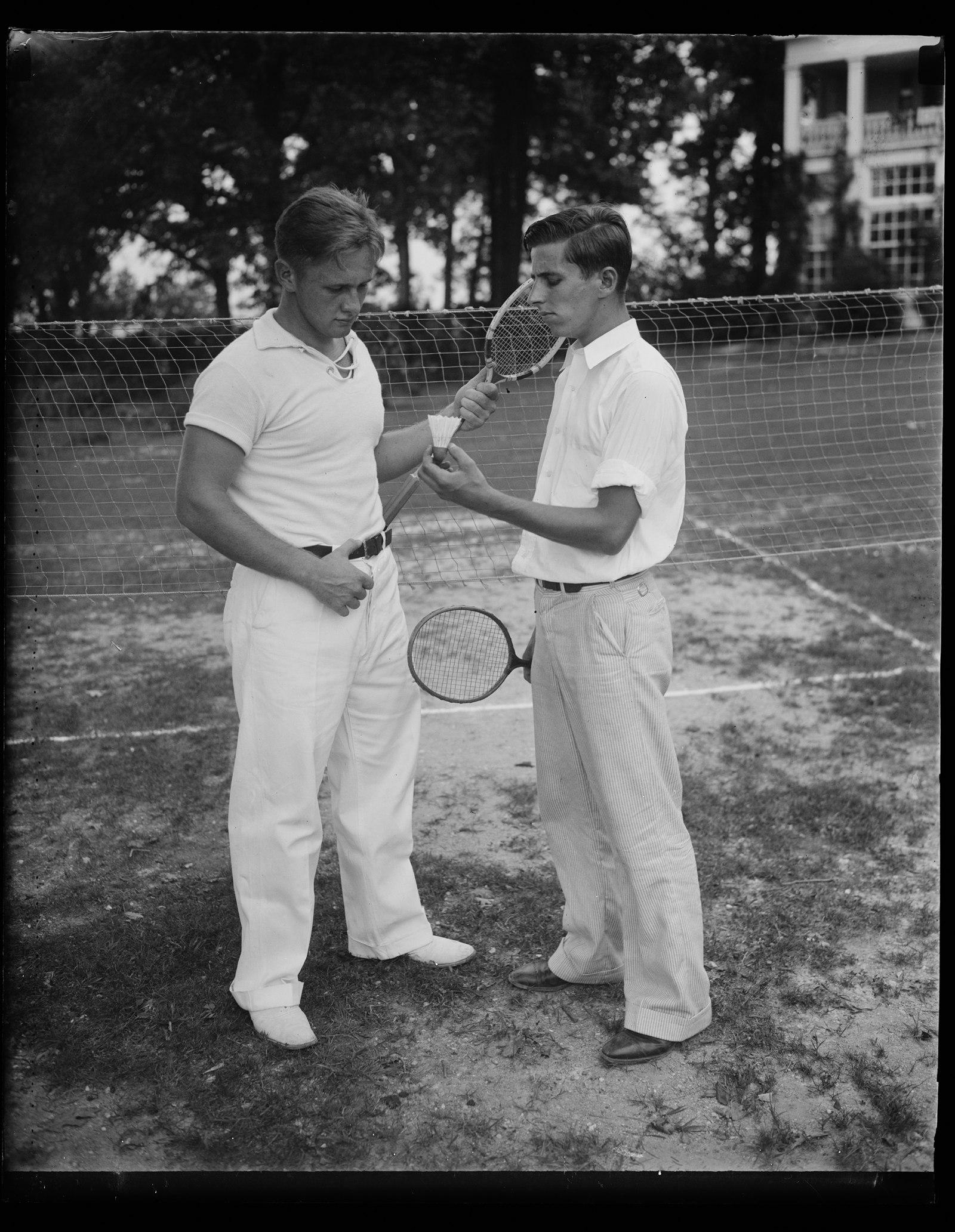 Two young men on a grass badminton court in 1936, one holding a shuttlecock, rackets in hand, the net strung behind them